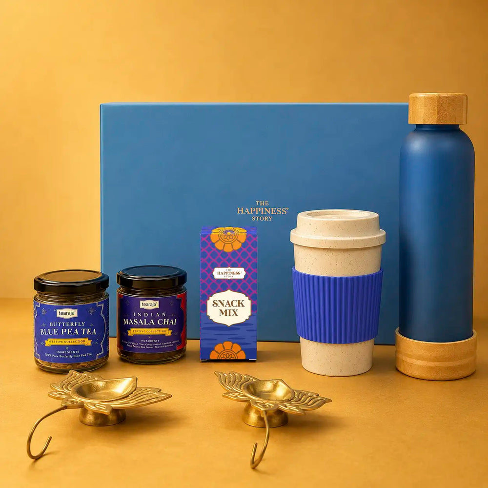 Diwali tea gift hamper contents set out in front of an ocean blue gift box — a frosted glass bottle with a bamboo cap, a wheat straw sipper mug with a blue grip, jars of butterfly blue pea tea and Indian masala chai, a snack mix carton and two brass lotus diya holders