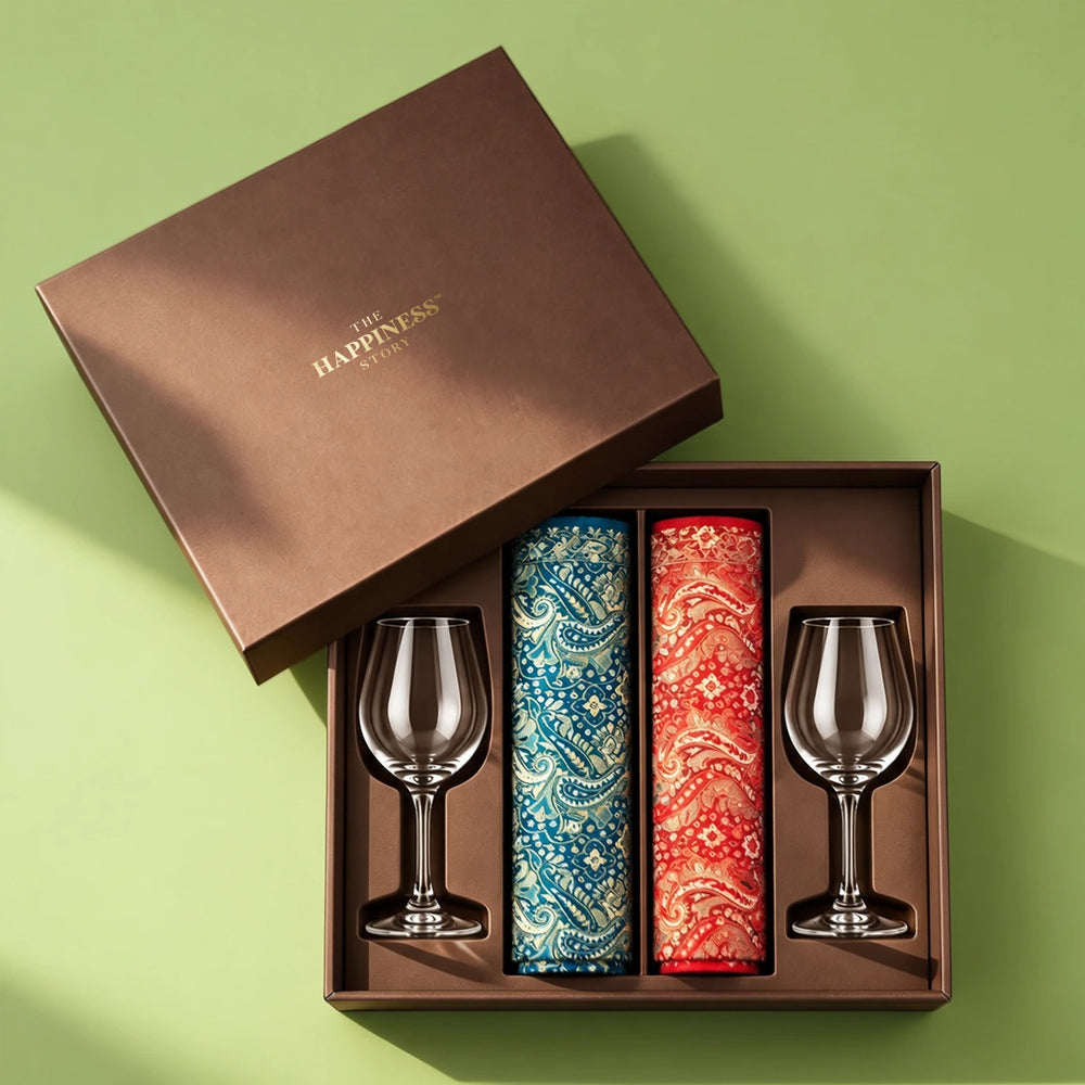 Open dusky brown gift hamper box with a gold foil lid, holding two rolled paisley jamawar shawls in blue and red and two clear stemmed wine glasses seated in a fitted tray