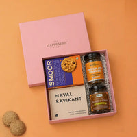 Open pink gift box with its lid lifted off, holding The Almanack of Naval Ravikant paperback, a jar of chocolate orange instant coffee, a jar of tulsi kadha chai and a carton of New York style chocolate chip cookies on white paper shred