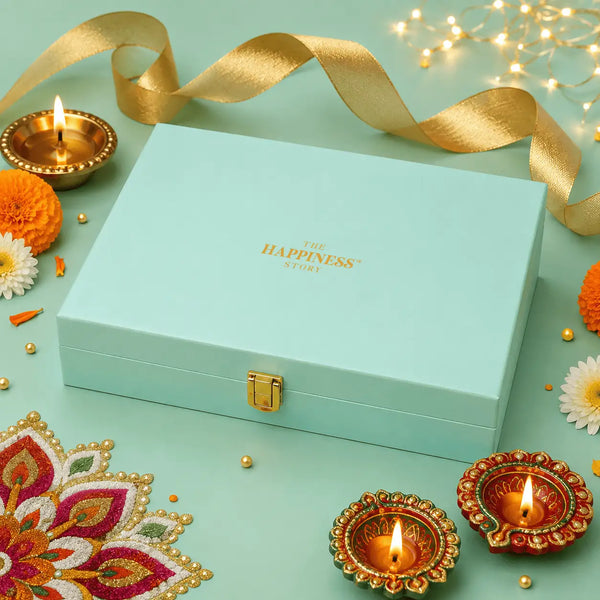 Closed mint green gift box with a gold metal clasp and a gold foil logo, surrounded by lit brass diyas, marigolds and a rangoli mat