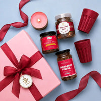 Flat lay of the tea and coffee gift hamper with the candle lit — pink ribboned gift box with its wish tag, rose green tea, cranberry instant coffee, coffee walnut cookies and two rice husk kullads