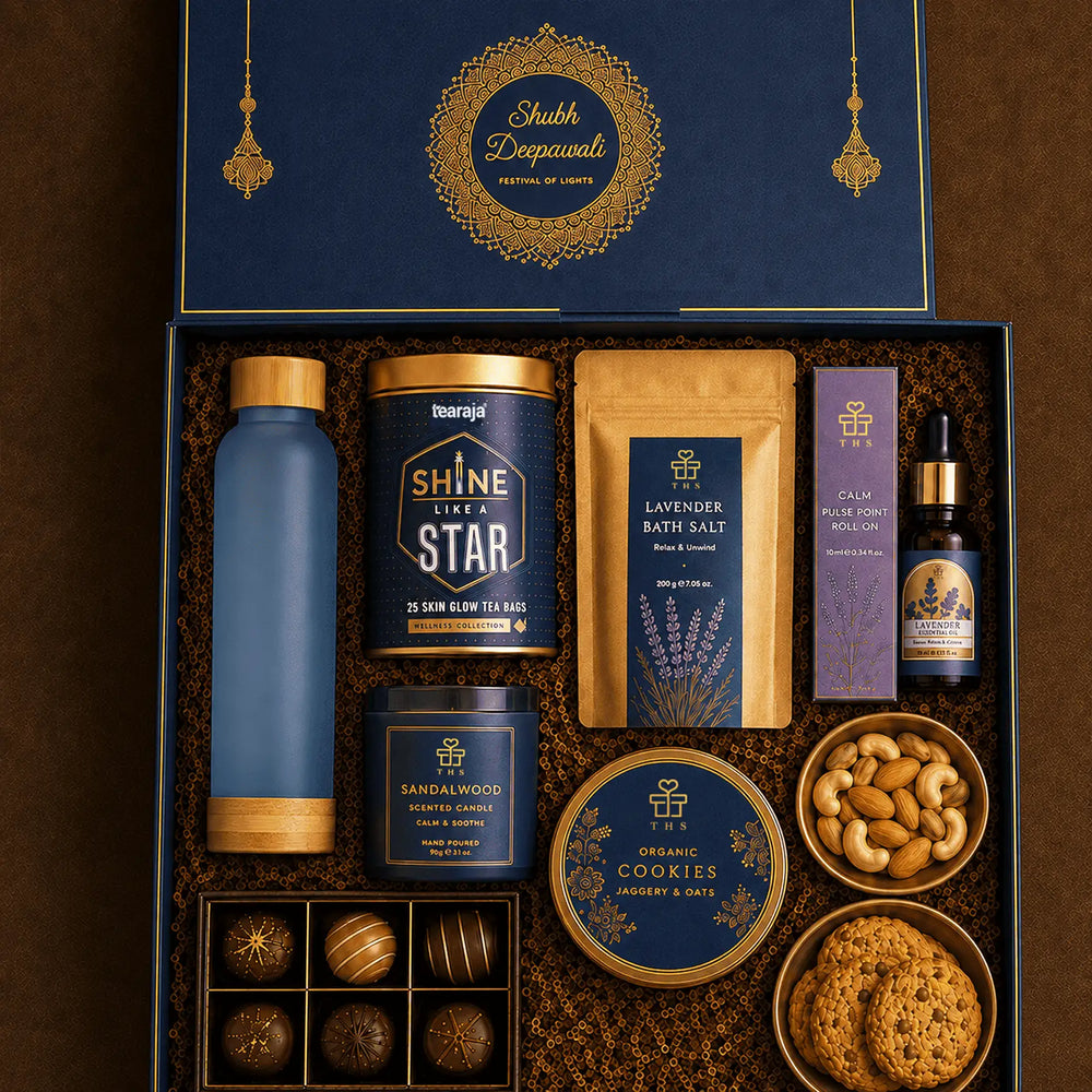 Open navy Diwali wellness gift box with a gold Shubh Deepawali mandala inside the lid, holding a frosted glass bottle, a tea tin, lavender bath salt, a pulse point roller, lavender essential oil, a sandalwood candle, cookies, dry fruits and six chocolates