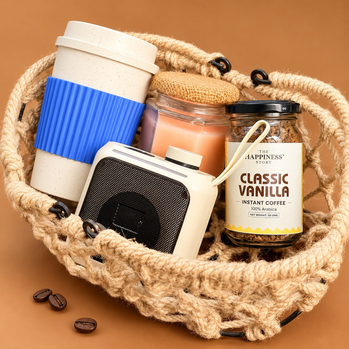 Employee onboarding welcome kit packed in a round jute rope basket with a cream bamboo fibre travel cup with a blue silicone grip, a cream portable Bluetooth speaker, a peach scented candle in a hexagonal glass jar with a jute lid, and a jar of Classic Vanilla instant coffee