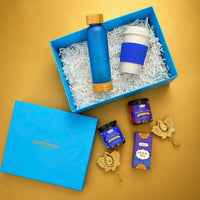 Ocean blue Diwali gift box seen from above with a frosted glass bottle and a wheat straw sipper mug inside, and jars of butterfly blue pea tea and Indian masala chai, a snack mix carton and two brass lotus diya holders laid out beside it