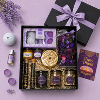 Open black Diwali gift box seen from above with a lavender satin bow, holding a lavender vaporizer set, scented candle, potpourri, dry fruit jars, mishri, mouth freshener, two tea tubes and a Happy Diwali card