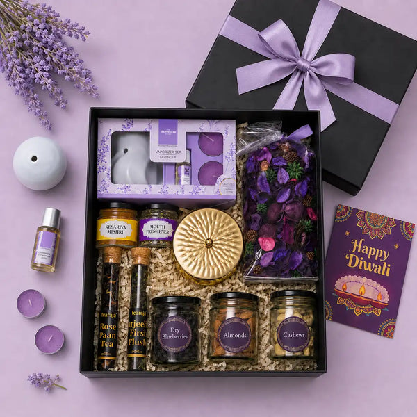 Open black Diwali gift box seen from above with a lavender satin bow, holding a lavender vaporizer set, scented candle, potpourri, dry fruit jars, mishri, mouth freshener, two tea tubes and a Happy Diwali card