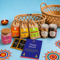 Contents of the Diwali gift basket laid out — three jute potlis of almonds, cashews and pistachios, jars of almond and coffee walnut cookies, a pink jasmine candle with a woven jute cap, two roasted chai sachets and a Happy Diwali card