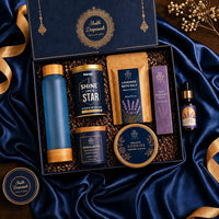 Diwali wellness hamper contents arranged in front of the closed navy gift box with marigolds and navy satin — frosted glass bottle, tea tin, lavender bath salt pouch, pulse point roller, essential oil, sandalwood candle, cookie and dry fruit tins and a box of chocolates