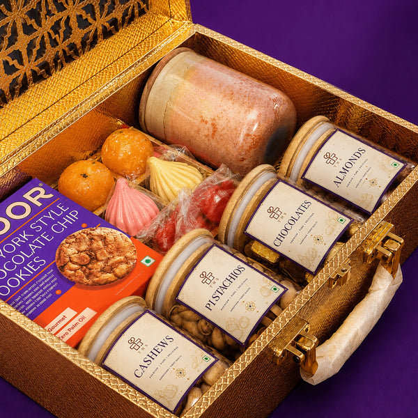 Close view inside the gold Diwali gift case showing four bamboo-lidded jars, a scented jar candle, a gold tray of modak and ladoo candles and a carton of chocolate chip cookies