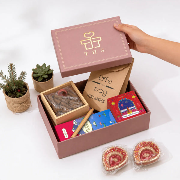 Open eco-friendly Diwali gift box with the lid lifted, showing rose dhoop cones in a bamboo-lidded box, incense sticks, a bamboo pen, a mini garden planter kit and two hand-painted diyas in crimson shred