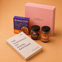Contents of the book gift hamper set out in front of the closed pink box with its gold-foil logo — paperback, cookie carton, tulsi kadha chai jar and chocolate orange coffee jar