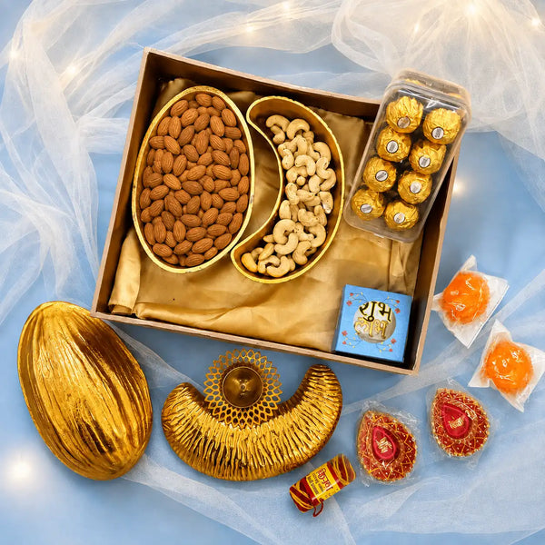 Luxury Diwali gift box with an almond-shaped gold platter of almonds and a cashew-shaped gold platter of cashews, beside a second pair of empty platters, a brass diya, painted diyas, ladoo candles, roli mouli, chocolates and a Shubh Labh