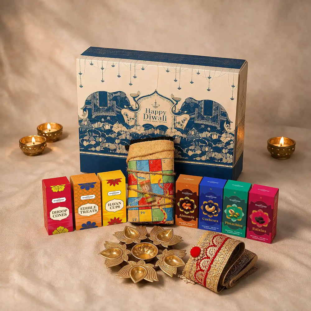 Eco-friendly Diwali gift hamper with a rolled jute ludo and snakes and ladders board, gold lotus urli, four dry fruit boxes, havan cups, dhoop cones and a gold-zari jute toran in front of a blue elephant-print keepsake gift box with lit diyas