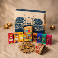 Eco-friendly Diwali gift hamper with a rolled jute ludo and snakes and ladders board, gold lotus urli, four dry fruit boxes, havan cups, dhoop cones and a gold-zari jute toran in front of a blue elephant-print keepsake gift box with lit diyas