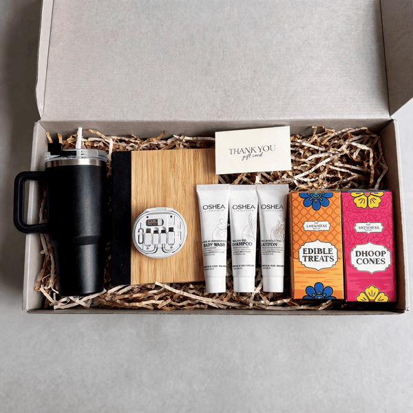 Flat overhead view of the open all-season gift hamper with the travel tumbler, bamboo notebook, cable kit, toiletries and treat cartons seated in shred filler