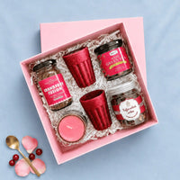 Rigid pink kappa board gift box with its lift-off lid to one side, packed with coffee and tea jars, a cookie jar, two rice husk kullads and a candle on white paper shred