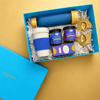 Open ocean blue Diwali gift box seen from above at an angle, holding a blue glass bottle, a wheat straw sipper mug with a blue grip, jars of masala chai and butterfly blue pea tea, a snack mix carton and two brass lotus diya holders