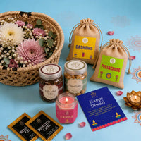 Close view of the Diwali gift basket contents — lit jasmine candle, jars of almond and coffee walnut cookies, jute potlis of cashews and pistachios, roasted chai sachets and a Happy Diwali greeting card