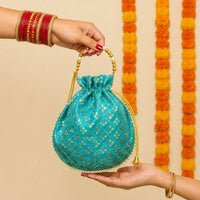 Woman's hands holding the turquoise Diwali potli gift bag by its gold beaded handle, showing it is about a hand span wide, marigold garlands behind