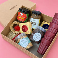 Open kraft Diwali gift box seen from the side, showing Kashmiri kahwa chai, vanilla instant coffee, almond and double chocolate cookie jars, two boxed ceramic diyas and maroon rice husk kulhads
