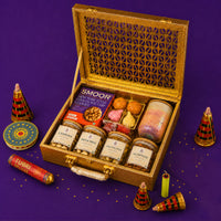 Gold Diwali gift case open at an angle, packed with jars of cashews, pistachios, chocolates and almonds, a scented candle, modak candles and chocolate chip cookies