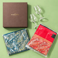 Blue and red paisley jamawar shawls folded flat beside a closed dusky brown gift box and two clear wine glasses, showing both shawl designs side by side