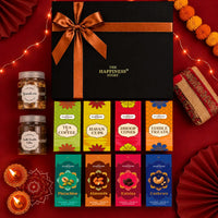 Overhead view of the black Diwali gift hamper contents: two jars of granola and double chocolate cookies, eight printed cartons of dry fruits and puja items, and a rolled gold-zari jute toran