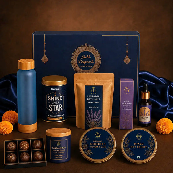Close view of the Diwali wellness hamper contents — a teal frosted glass bottle with a bamboo cap, a skin glow tea tin, a kraft lavender bath salt pouch, a lavender pulse point roller carton, an amber essential oil dropper bottle, a sandalwood candle and two navy and gold tins