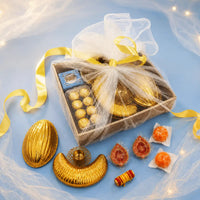 Luxury Diwali gift box wrapped in white net with a yellow satin ribbon, with the gold almond and cashew platters, brass lotus diya, hand-painted diyas, ladoo candles and roli mouli set out in front