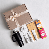 Closed kraft gift box with a satin ribbon beside the full hamper contents laid out: travel tumbler, bamboo notebook, utility kit, toiletries and treat cartons