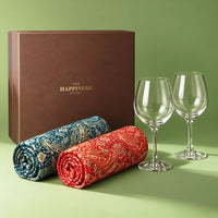 Closed dusky brown keepsake gift box with a gold foil wordmark, with two rolled jamawar paisley shawls in teal blue and scarlet and a pair of wine glasses standing beside it