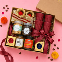 Overhead view of an open Diwali gift hamper showing a jute toran, four rice husk kulhad cups, almond cookies, double chocolate cookies, vanilla instant coffee, Kashmiri kahwa chai and two boxed ceramic diyas in a kraft tray