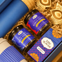 Close view inside the Diwali tea hamper showing the bamboo cap of the ocean blue glass bottle, jars of Indian masala chai and butterfly blue pea tea, a snack mix carton and a brass lotus diya holder