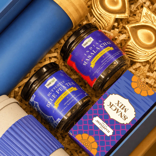 Close view inside the Diwali tea hamper showing the bamboo cap of the ocean blue glass bottle, jars of Indian masala chai and butterfly blue pea tea, a snack mix carton and a brass lotus diya holder