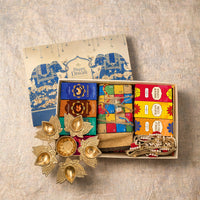 Open Diwali gift hamper packed in its tray with the printed lid beside it, showing four dry fruit cartons, the twine-tied jute ludo board with dice, three festive cartons, a coiled gold-zari jute toran and a lotus urli