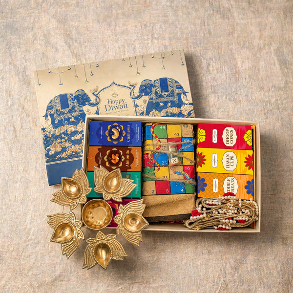 Open Diwali gift hamper packed in its tray with the printed lid beside it, showing four dry fruit cartons, the twine-tied jute ludo board with dice, three festive cartons, a coiled gold-zari jute toran and a lotus urli