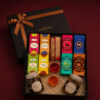Open black kappa board Diwali gift box with its ribboned lid behind, packed with printed dry fruit and puja cartons, a rolled jute toran and jars of granola and cookies in cream shred filler