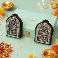 Shubh and Labh plaques in embossed antique silver-finish metal on dark wooden bases, standing either side of a mint green gift box with a gold foil logo
