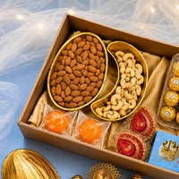 Open Diwali hamper box holding a gold almond-shaped platter filled with almonds and a gold cashew-shaped platter filled with cashews, with ladoo candles, hand-painted diyas, foil-wrapped chocolates and a Shubh Labh box