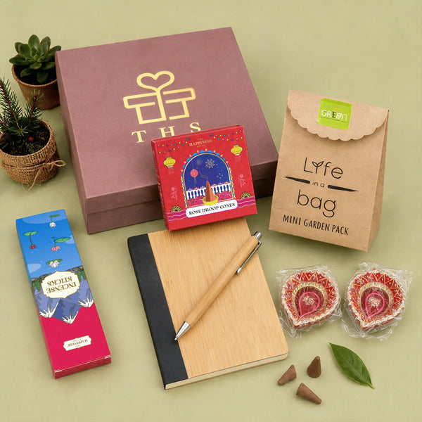 Eco-friendly Diwali gift hamper contents — bamboo notebook and bamboo pen, rose dhoop cones box, incense sticks box, mini planter grow kit and two hand-painted earthen diyas beside a dusty rose gift box