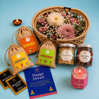 Diwali gift basket contents with the jasmine candle lit in its hexagonal glass jar, beside jute potlis of dry fruits, two cookie jars, roasted chai sachets and a Happy Diwali card