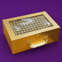 Closed gold Diwali gift case with a carry handle, two gold clasps and a laser-cut lattice panel set behind a clear window in the lid