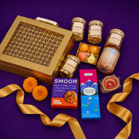 Gold Diwali gift case at an angle with jars of dry fruits and chocolates, a scented candle, incense sticks, modak candles and a hand-painted diya arranged beside a gold ribbon