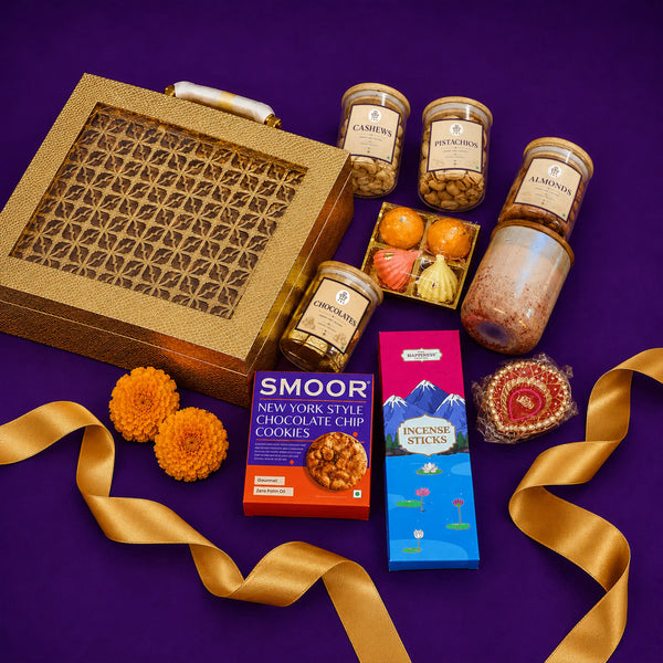 Gold Diwali gift case at an angle with jars of dry fruits and chocolates, a scented candle, incense sticks, modak candles and a hand-painted diya arranged beside a gold ribbon