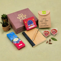 Dusty rose kappa board Diwali gift box with gold foil logo, shown with a bamboo notebook and pen, rose dhoop cones, incense sticks, a planter grow kit and two hand-painted diyas