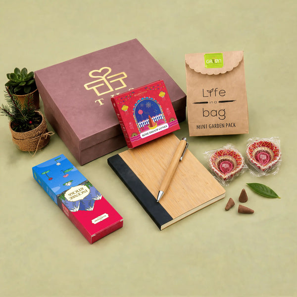 Dusty rose kappa board Diwali gift box with gold foil logo, shown with a bamboo notebook and pen, rose dhoop cones, incense sticks, a planter grow kit and two hand-painted diyas