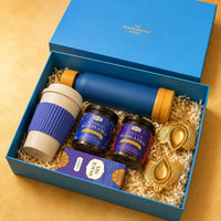 Ocean blue Diwali gift box with its lid standing behind, packed in a single layer with a frosted glass bottle, a wheat straw sipper mug, two loose leaf tea jars, a snack mix carton and two brass lotus diya holders
