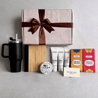 Closed gift box with the hamper contents lined up in front: travel tumbler, bamboo notebook, utility travel kit, toiletries, chocolates and dhoop cones