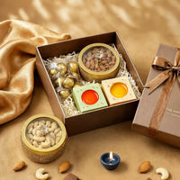 Open Diwali hamper box with a gold cashew jar lifted out beside it, two ceramic diyas and a pouch of foil-wrapped chocolates inside