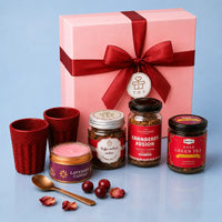 Pink gift box tied with a deep red ribbon and a round wish tag, with two rice husk kullads, a lavender candle and jars of cranberry coffee, coffee walnut cookies and rose green tea set out in front